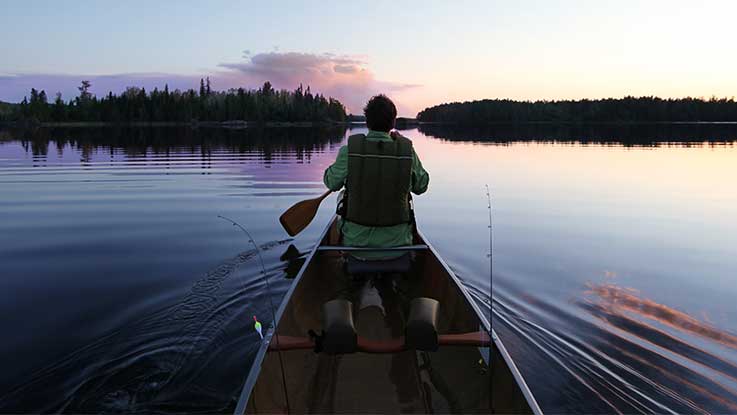 Ein POV vom Rücksitz eines Kanus aus, wie ein Mann auf dem Vordersitz des Kanus an einem ruhigen Sommerabend auf einem ruhigen See paddelt.