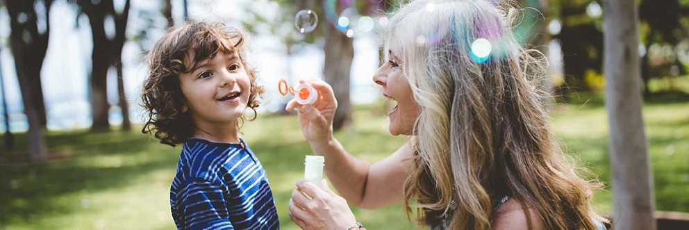 A grandmother kneels down to blow bubbles with her grandson in the park.