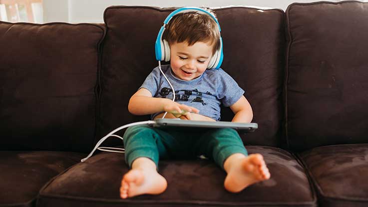 A toddler sitting in the middle of a couch smiles happily as he plays on a tablet. 