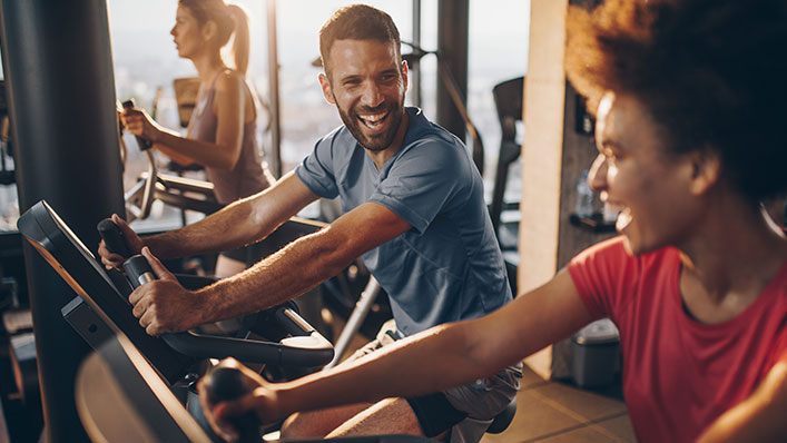 Man and woman laughing and chatting while riding exercise bikes in a gym.