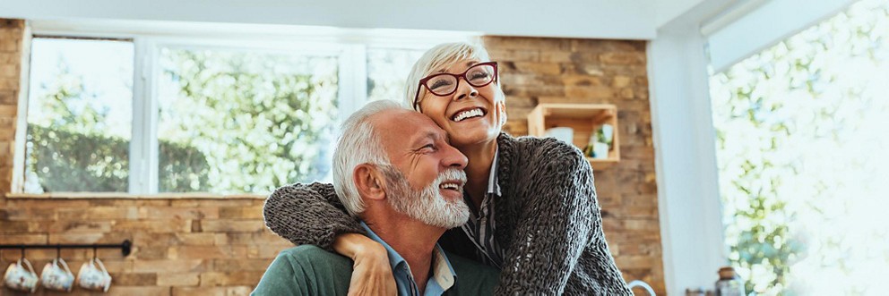 An older couple laughs as they embrace in a bright kitchen.