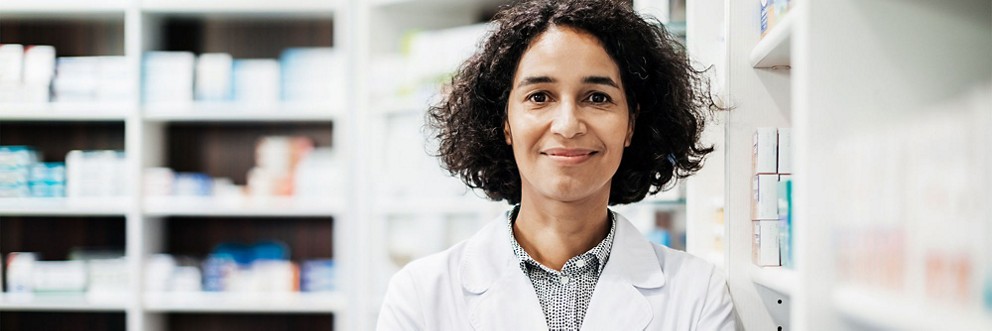 A pharmacist standing in front of medicine shelves smiles at the camera.