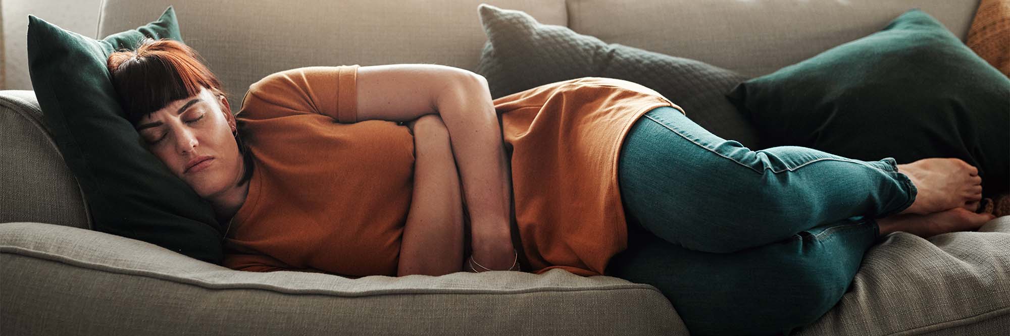  A woman lays on a couch frowning with her arms wrapped around her lower stomach
