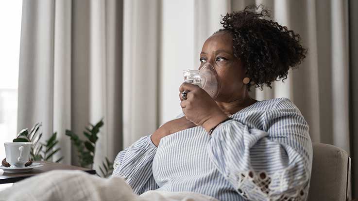 A woman at home breathes deeply from the oxygen mask she holds onto her face.