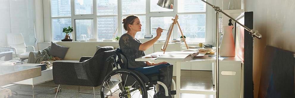 A woman in a wheelchair uses an easel to paint in her bright apartment.