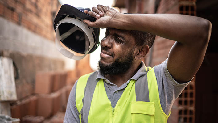 A male construction worker removes his hard hat to wipe the sweat from his brow.