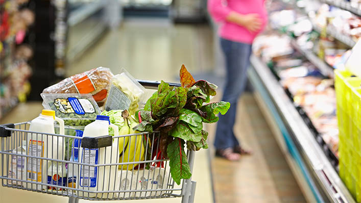 A pregnant woman browses the aisles at the grocery store with her cart full of lettuce, bananas, bread, and milk.