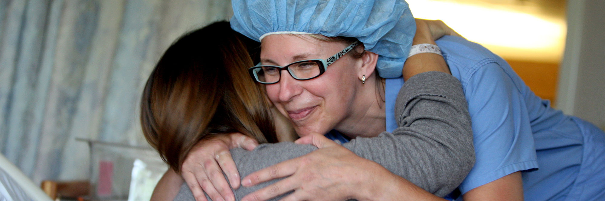 A smiling nurse hugs a patient in a hospital bed.