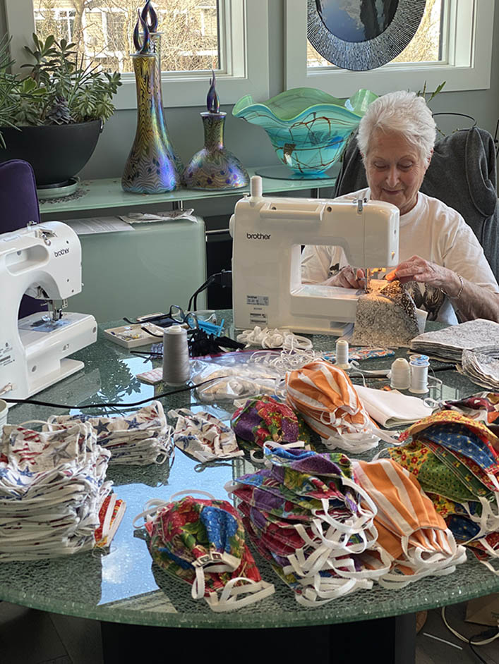 Donna sits at the table with her sewing machine and stacks of fabric masks.