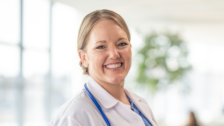 A headshot of a blonde, female doctor. She smiles at the camera and wears a white coat and stethoscope.